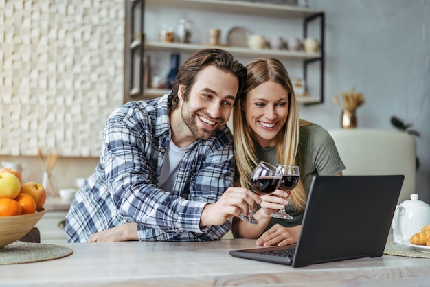 Happy young european man with stubble and lady clink glasses with wine and look at laptop in modern