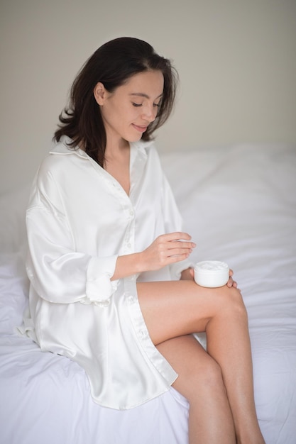 Happy young european woman in bathrobe with perfect legs sits on white bed with jar of cream in bedroom