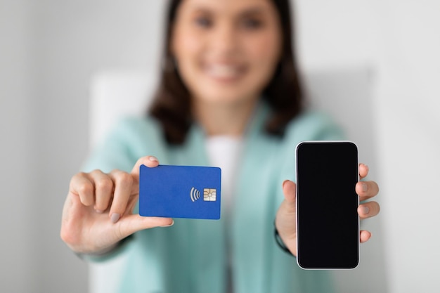 Happy young european woman in suit shows smartphone with blank screen recommends credit card