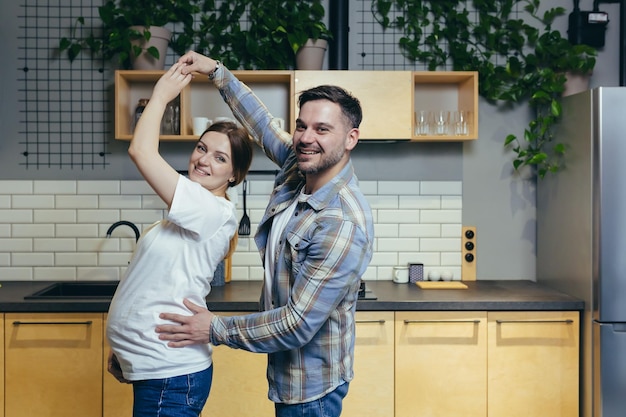 Happy young family in anticipation of a child dancing in the kitchen together looking at the camera smiling Pregnant woman and man