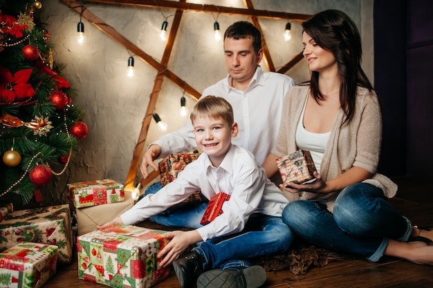 Happy young family in Christmas decorations, mom, dad and little boy near Christmas tree with presents near