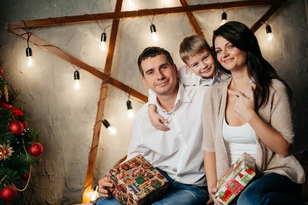 Happy young family in Christmas decorations, mom, dad and little boy near Christmas tree with presents near