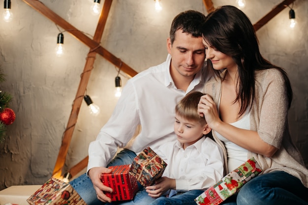 Счастливая молодая семья в рождественских украшениях, мама, папа и маленький мальчик возле елки с подарками рядом