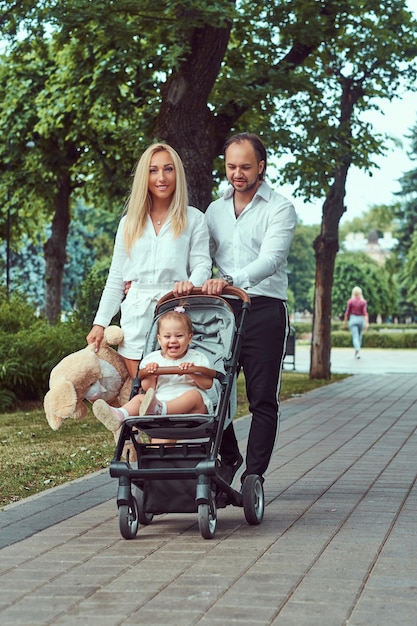 幸せな若い家族の概念。美しいブロンドの母親とひげを生やしたスタイリッシュな父親は、ベビーカーで小さな娘と一緒に公園を歩いています。