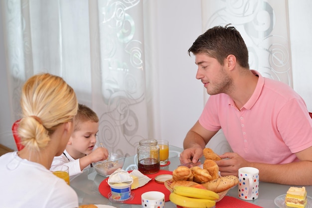 幸せな若い家族は、明るい朝の光に赤の詳細とキッチンで健康的な朝食を持っています。