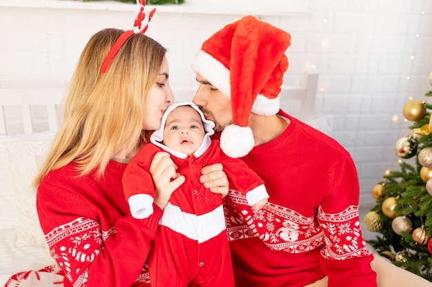 Happy young family mom dad and baby in red sweaters under the Christmas tree on the bed at home celebrate New Year or Christmas by kissing and hugging each other