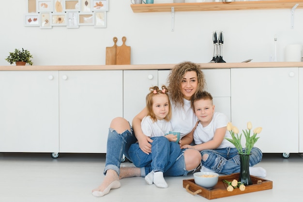 Happy young family, mom with little children sit on the floor in the kitchen