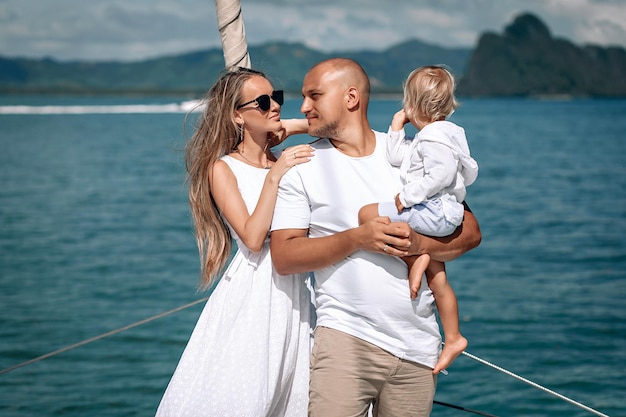 Foto felice giovane famiglia in abiti bianchi - donna bionda dai capelli lunghi, bambino e uomo calvo in piedi insieme sullo yacht. sfondo blu del mare. phuket. tailandia