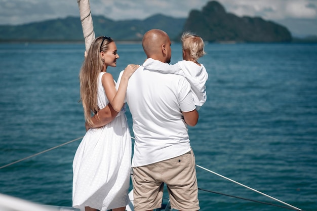 Foto felice giovane famiglia in abiti bianchi - donna bionda dai capelli lunghi, bambino piccolo e uomo calvo in piedi insieme sullo yacht e guardando il mare blu. phuket. tailandia