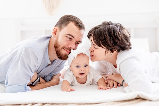 a happy young family with a newborn baby hugging and kissing on the bed at home in a bright room mom dad and a small child on a white bed smiling
