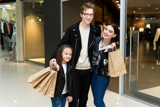 Счастливая молодая семья с бумажными сумками ходя по магазинам на моле. Магазинные витрины с одеждой