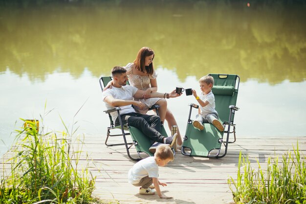 Happy young family with toddler children resting sitting on folding camping chairs over lake on a wooden pier outdoors. spend leisure time together in camp pontoon with kids in nature. vacation forest