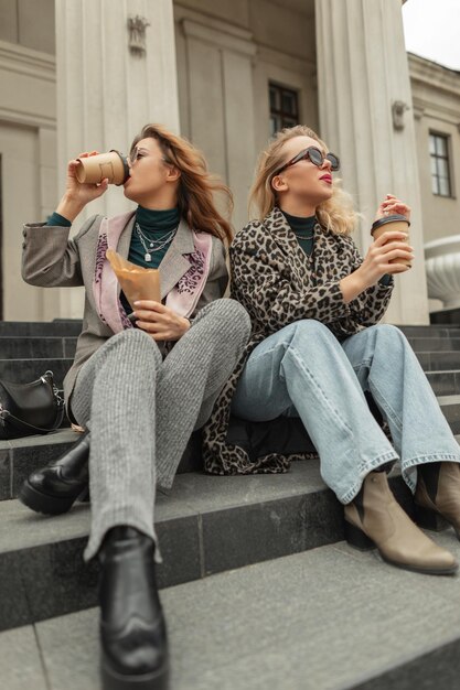 Фото Счастливые молодые модные красивые девушки в модной модной одежде сидят на ступеньках, пьют кофе и едят профитроли в городе. люди путешествуют и наслаждаются образом жизни