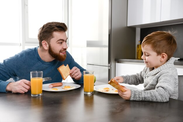 happy young father dressed in blue sweater eating at kitchen with his little son