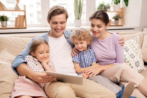 Happy young father holding tablet while his wife and two cute kids sitting next to him