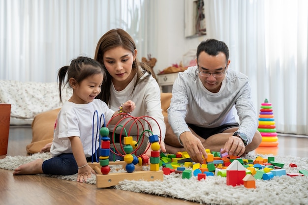 幸せな若い父と母と小さな娘がおもちゃの木製ブロックで遊んで、リビングルーム、家族、親子関係、発達のおもちゃで人々の概念の床に座っています