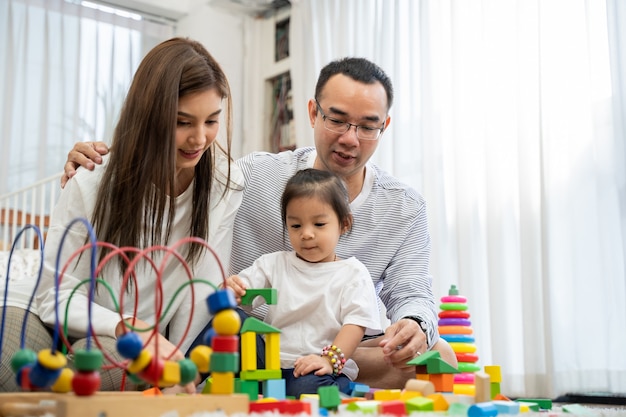 幸せな若い父と母と小さな娘がおもちゃの木製ブロックで遊んで、リビングルーム、家族、親子関係、発達のおもちゃで人々の概念の床に座っています