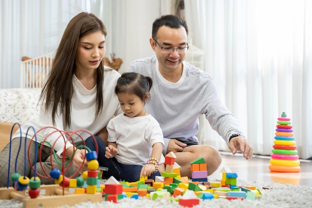 幸せな若い父と母と小さな娘がおもちゃの木製ブロックで遊んで、リビングルーム、家族、親子関係、発達のおもちゃで人々の概念の床に座っています