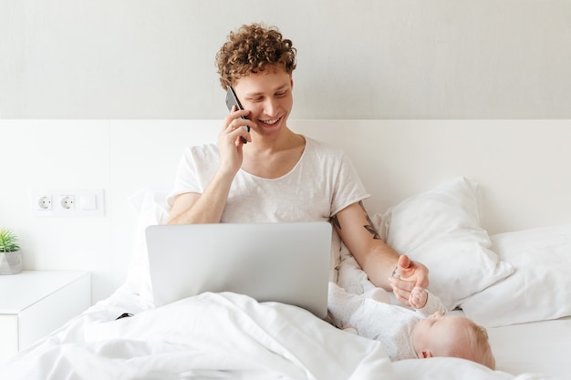 Happy young father playing with his little baby son while working on a laptop computer, laying in bed, talking on mobile phone