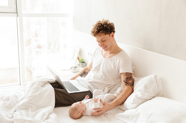 Happy young father playing with his little baby son while working on a laptop computer, laying in bed