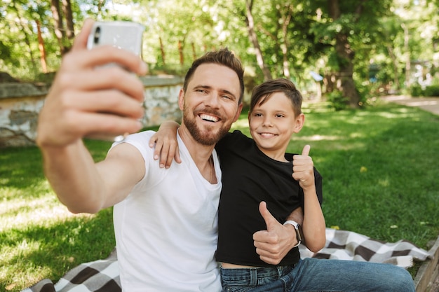 彼の幼い息子と座っている幸せな若い父親は携帯電話でselfieを作る