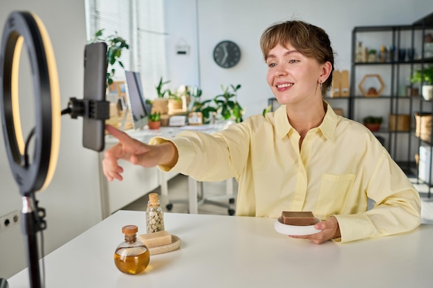 Happy young female blogger pointing at smartphone screen while pressing record button before making livestream about unpacking