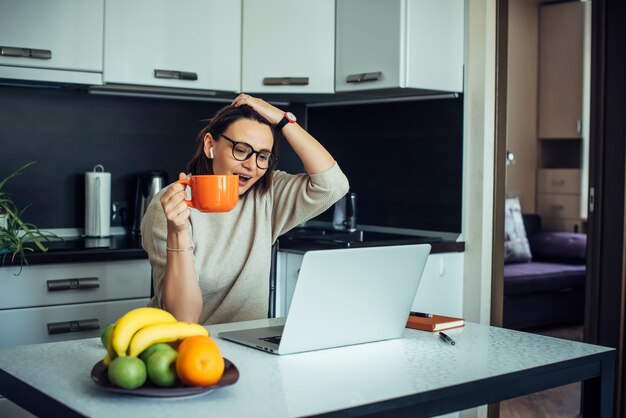 Happy young female blogger wearing glasses in home kitchen with laptop broadcasting live