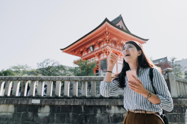 Happy young female college girl student tourist searching\
location on smartphone standing visiting in travel destination with\
red japanese temple in the background. kiyomizu dera shrine kyoto\
japan.