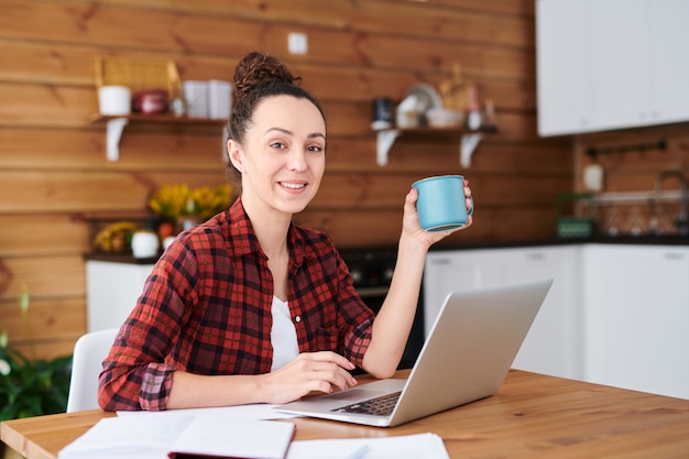 Счастливая молодая женщина-дизайнер или фрилансер с чашкой чая или кофе просматривает веб-сайты в поисках новых идей на кухне