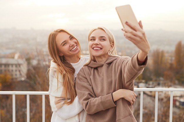 Giovani amiche felici in abiti caldi ed eleganti sorridenti e prendendo selfie sullo smartphone mentre si sta in piedi sul ponte di osservazione in città durante il fine settimana insieme