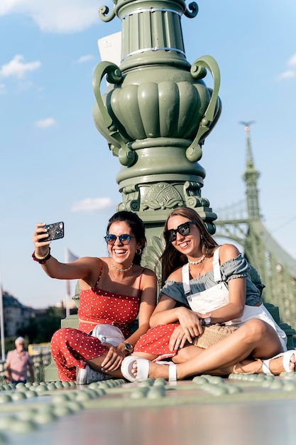 Happy young female friends in sunglasses sitting near column and browsing internet on mobile phone  of city street