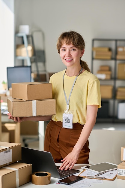 Happy young female owner of warehouse lookng at camera by workplace with laptop while holding stack of boxes and checking online data