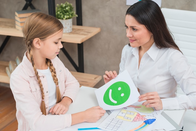 ブロンドの女の子に幸せな緑の感情顔カードを示す幸せな若い女性心理学者