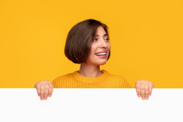 Happy young female smiling and looking away while demonstrating blank poster during promotion campaign against yellow background