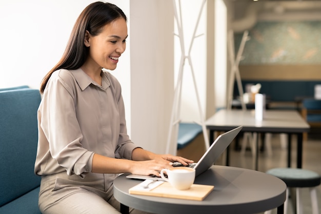 Happy young female student in smart casual sitting by table in cafe