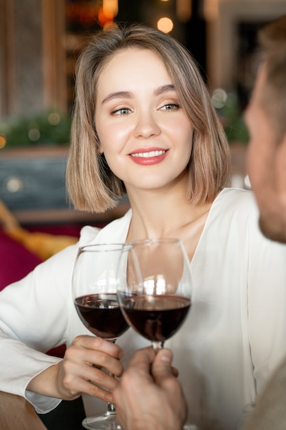 Happy young female with glass of red wine looking at her boyfriend with toothy smile during toast for their relationship in restaurant