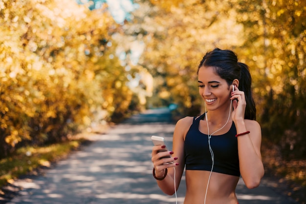 Happy young fit woman using cell phone while listening to music on smartphone in park.