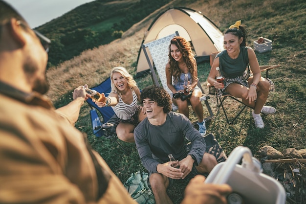 Happy young friends enjoy a sunny day at the mountain. They're laughing and drinking beer near tent.