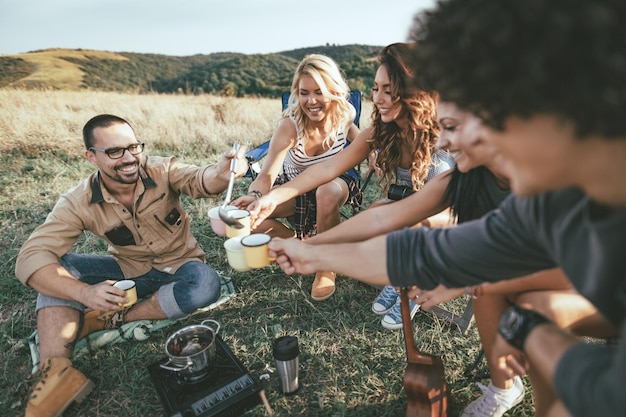 happy young friends enjoy a sunny day at the mountain. they're laughing and grilling -roasting sausages on sticks over a campfire near tent.