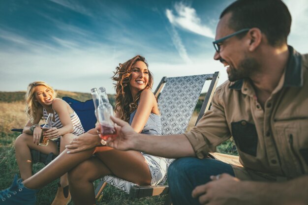 Happy young friends enjoy a sunny day at the mountain. They're laughing and toasting with beer bottles near tent.