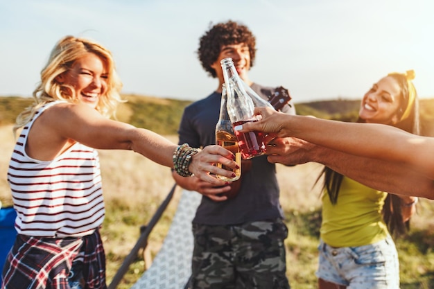 Happy young friends enjoy a sunny day in nature. They're clinking beer bottles, laughing and talking happy to be together.