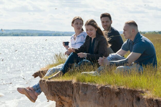 Happy young friends sitting on the hill over the river enjoying recreation and drinking tea