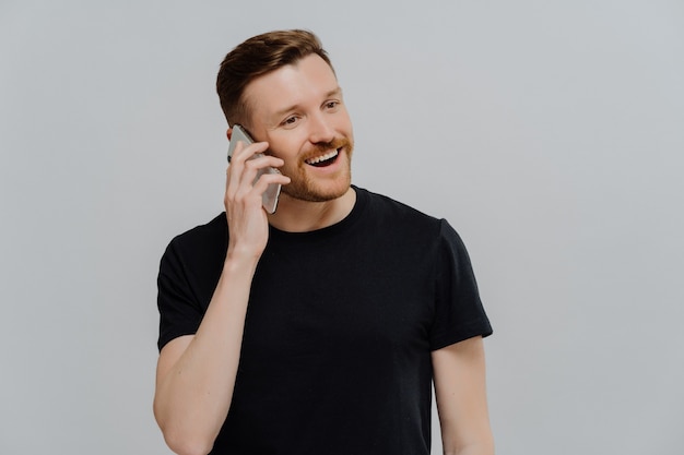 Happy young ginger man with stubble talking on mobile phone and feeling satisfied, receiving positive good news, guy with red hair looking aside and smiling while standing against grey studio wall