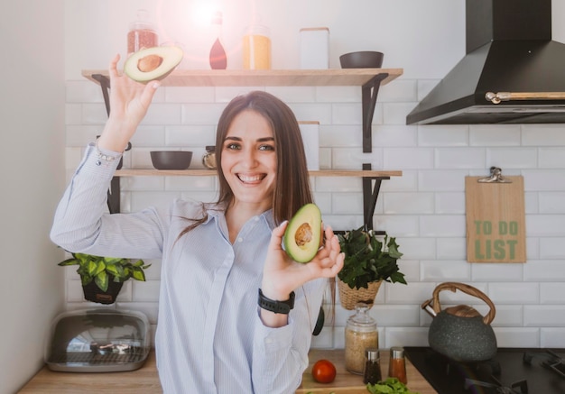 happy young girl holding cut avocado halves on kitchen background