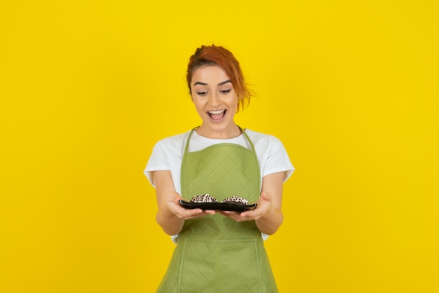 Happy young girl holding fresh homemade cookies on yellow wall