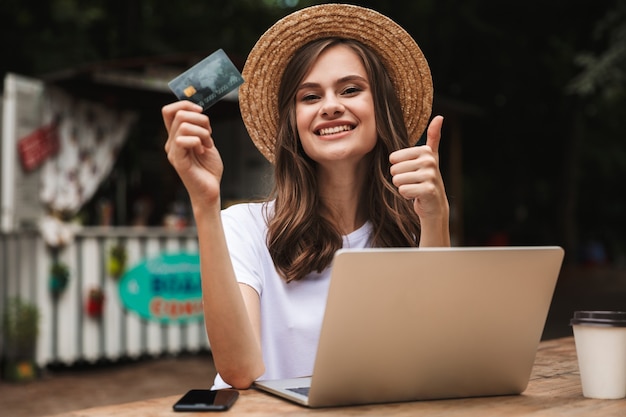 Happy young girl holding plastic credit card while sitting with\
laptop computer and coffee at the cafe outdoors, showing thumbs\
up