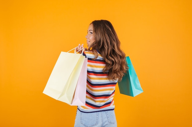 Happy young girl posing over yellow wall space holding shopping bags.