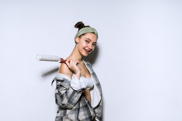 Happy young girl in a shirt paints a wall with a roller in her new apartment