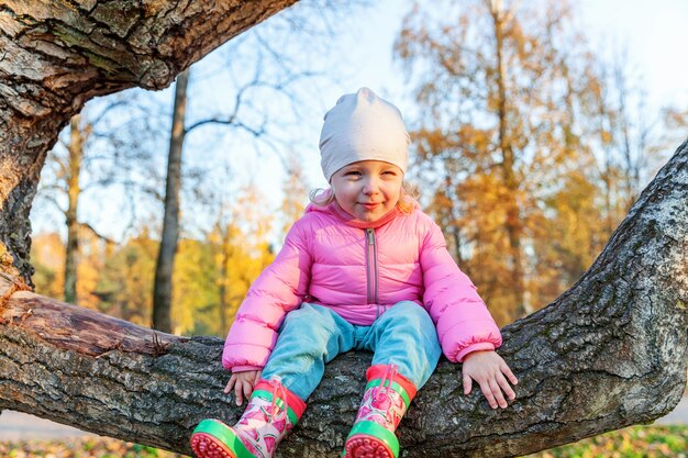 Happy young girl smiling and sitting on tree in beautiful autumn park on nature walks outdoors. Little child playing in autumn fall orange yellow background. Hello autumn concept.