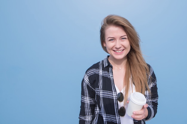 Happy young girl standing on a blue background with a cup of coffee in her hands smiles and looks at the camera